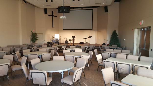 An empty church hall with chairs and tables facing a stage with a cross and musical equipment.