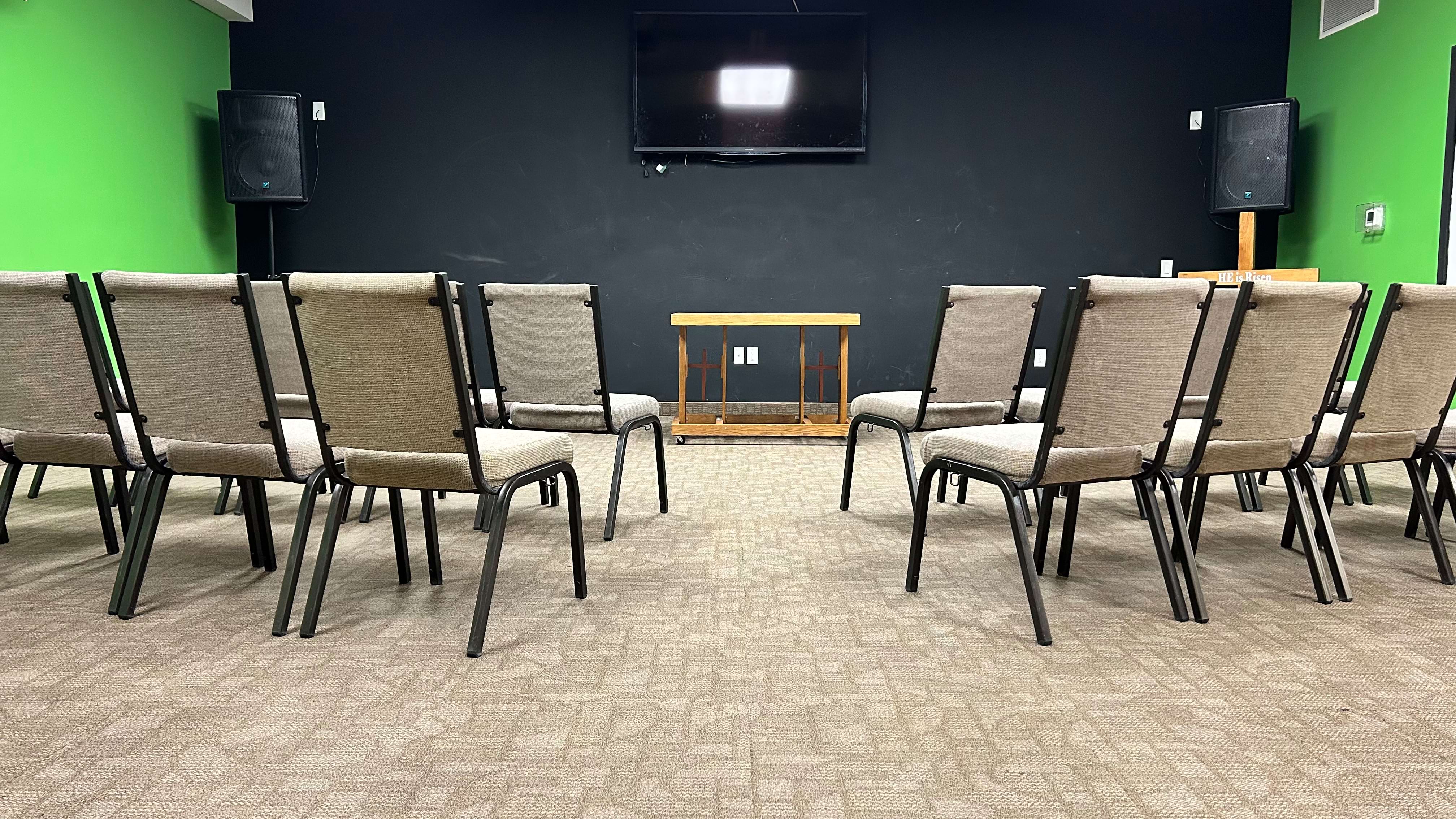 A row of empty chairs facing a wall-mounted TV and speakers in a room with green and black walls.