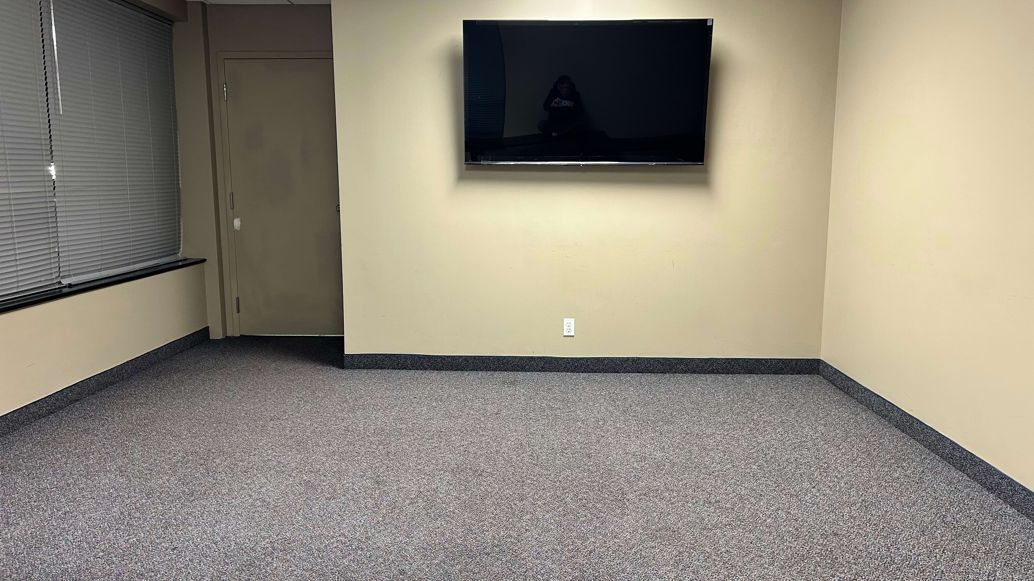 Empty room with beige walls, a mounted TV, window blinds on the left, and a closed door.