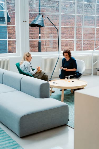 Two people sit and converse in a modern living room with large windows, a gray sofa, and a round coffee table.