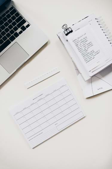 Laptop, notebook, planner, and pen on a white desk, suggesting a work or study environment.