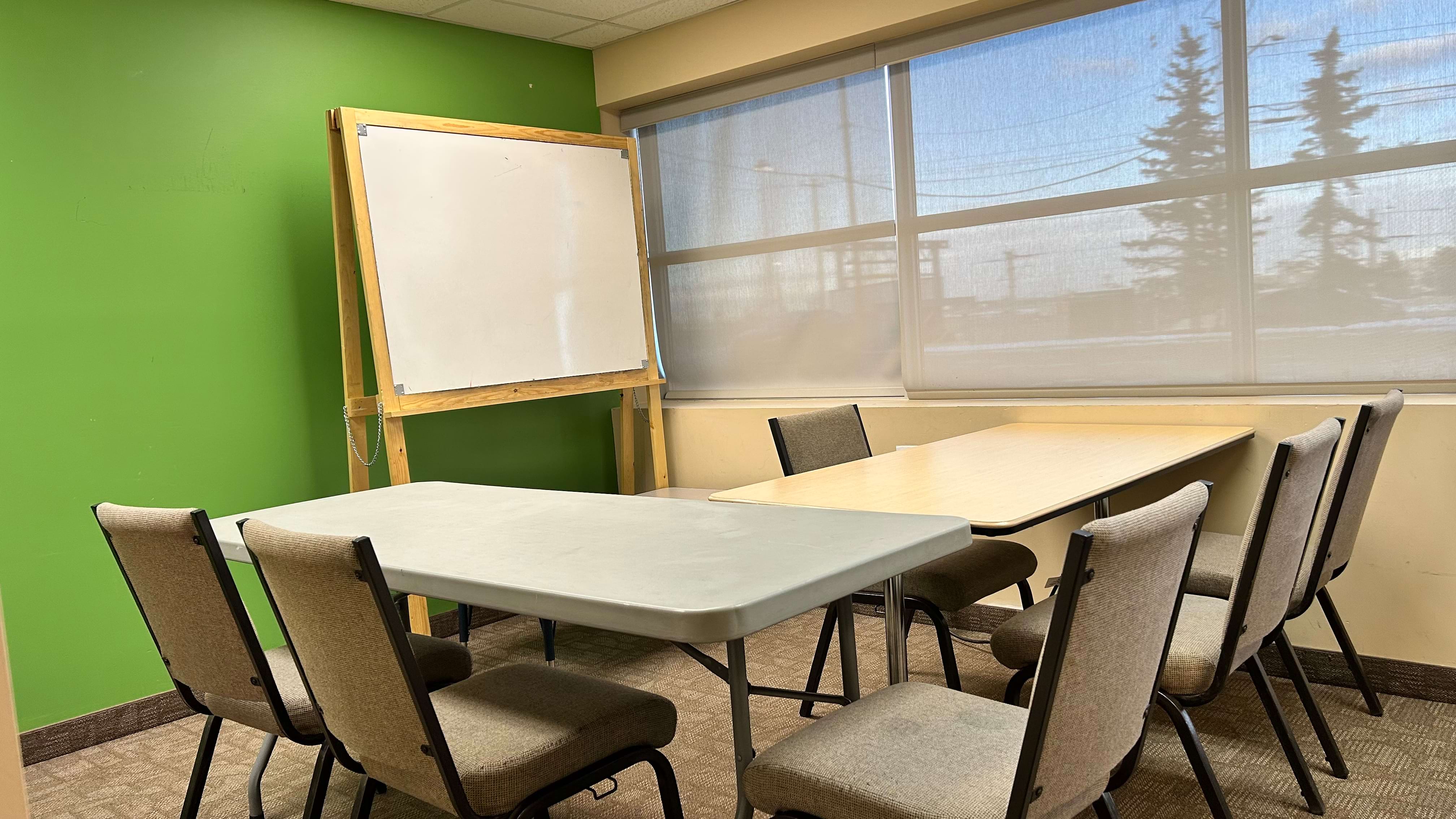 Meeting room with tables, chairs, whiteboard, and window with trees outside.