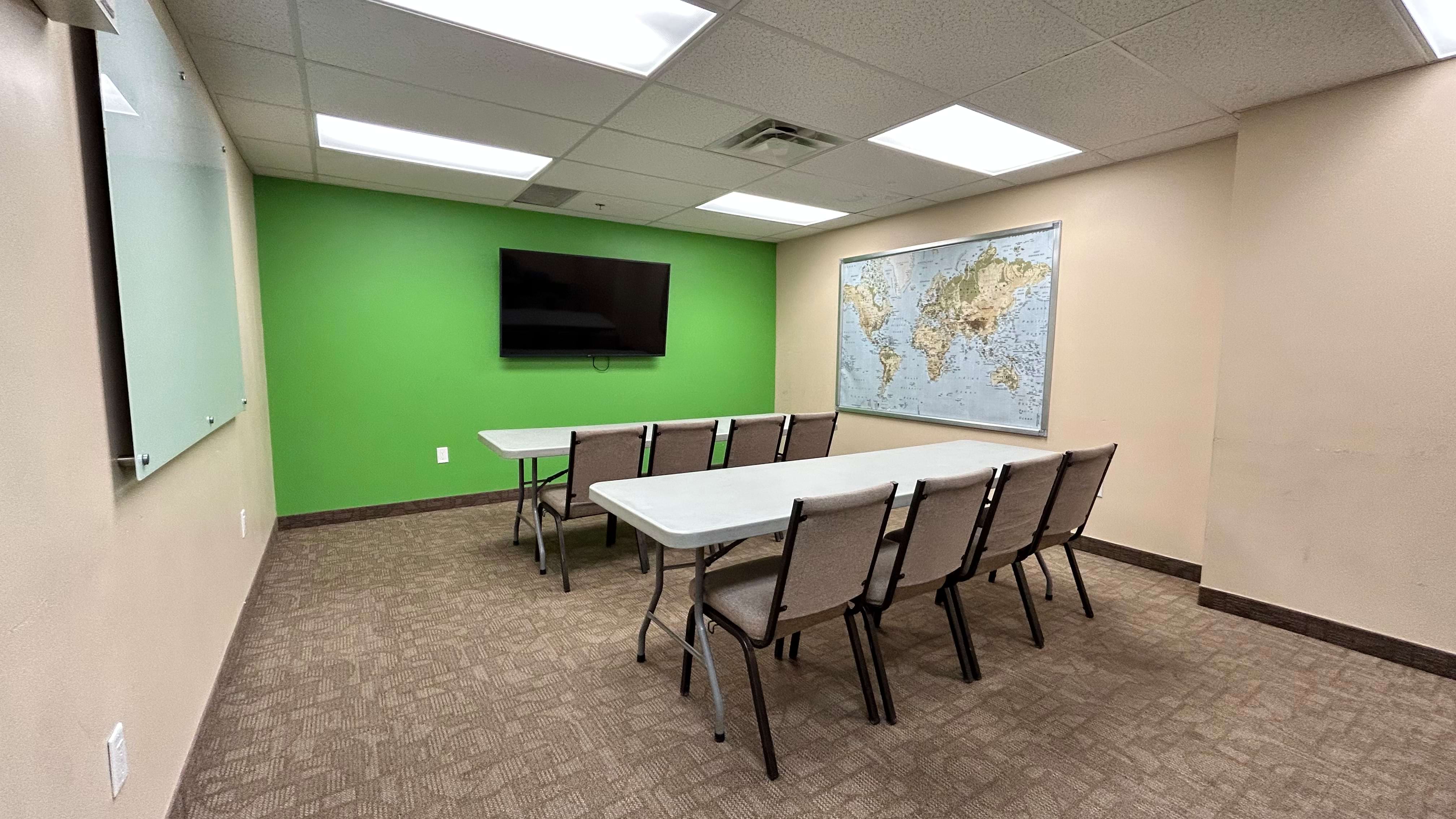 A minimalist meeting room with a green wall, world map, TV screen, and two white tables with chairs.